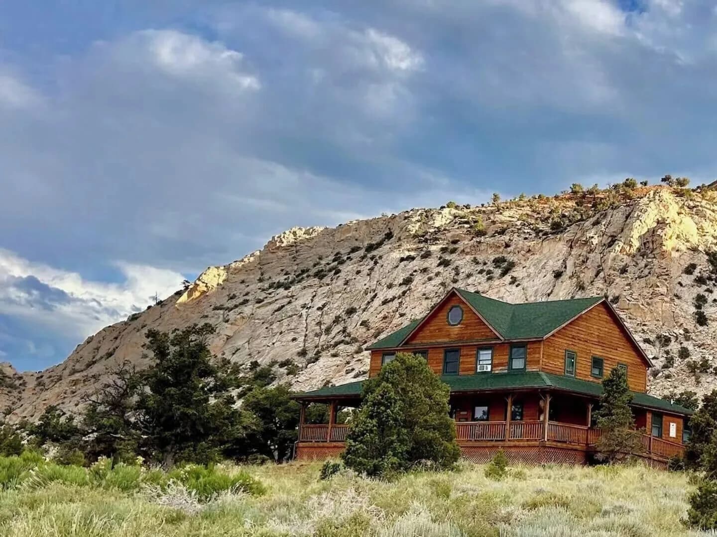 The Lodge seen from the sagebrush below, a two-storey timber building with a green roof and a wraparound covered deck, under a white sandstone ridge